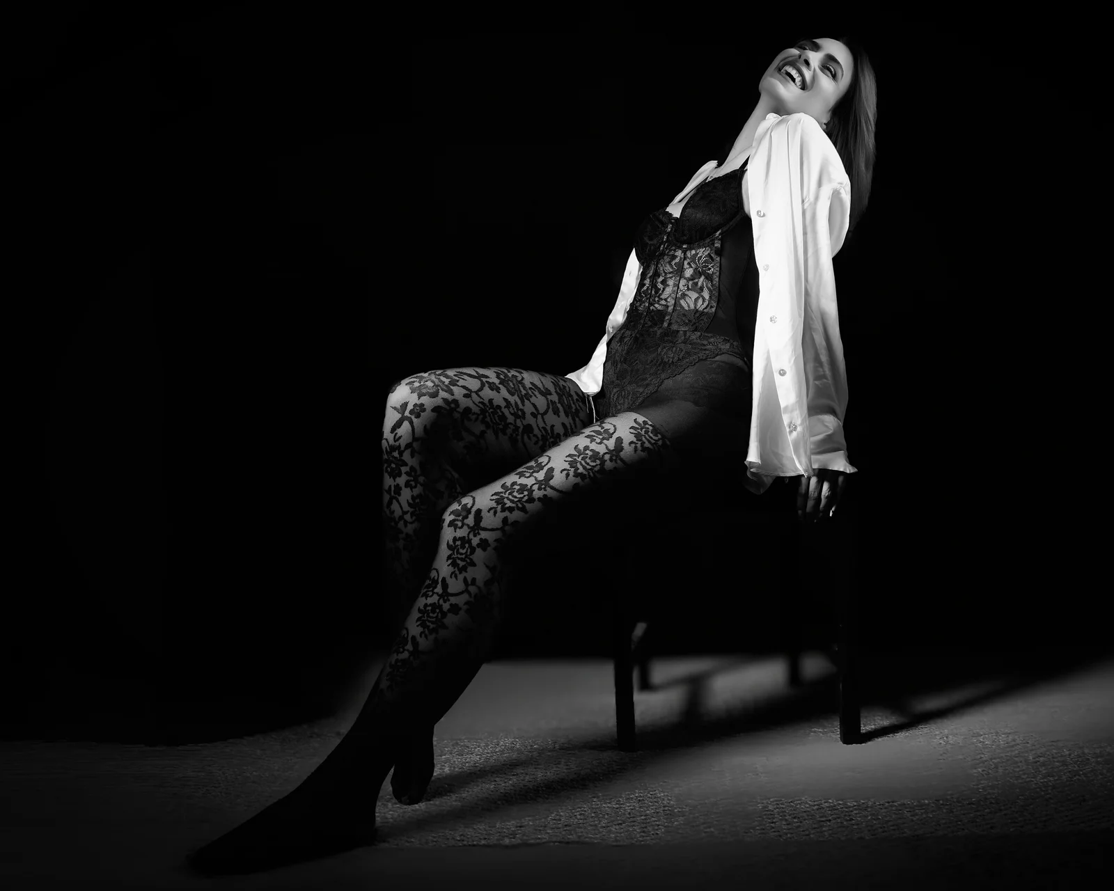 Femme riant tête en arrière, assise sur une chaise, en dentelle noire et chemise blanche ouverte, lumière douce, noir et blanc