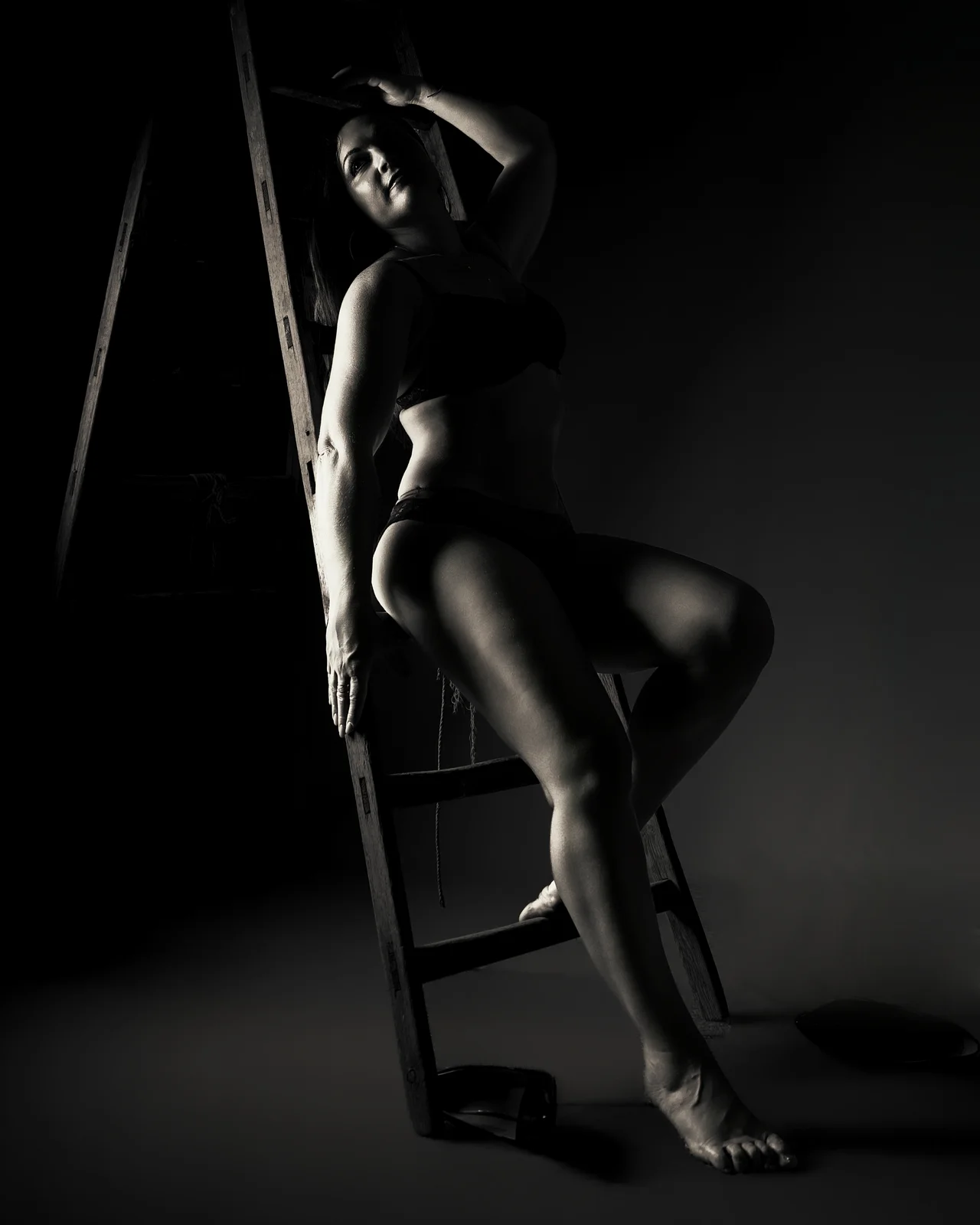 Femme en lingerie noire appuyée contre une vieille échelle en bois, regard levé, éclairage latéral en clair-obscur, fond sombre