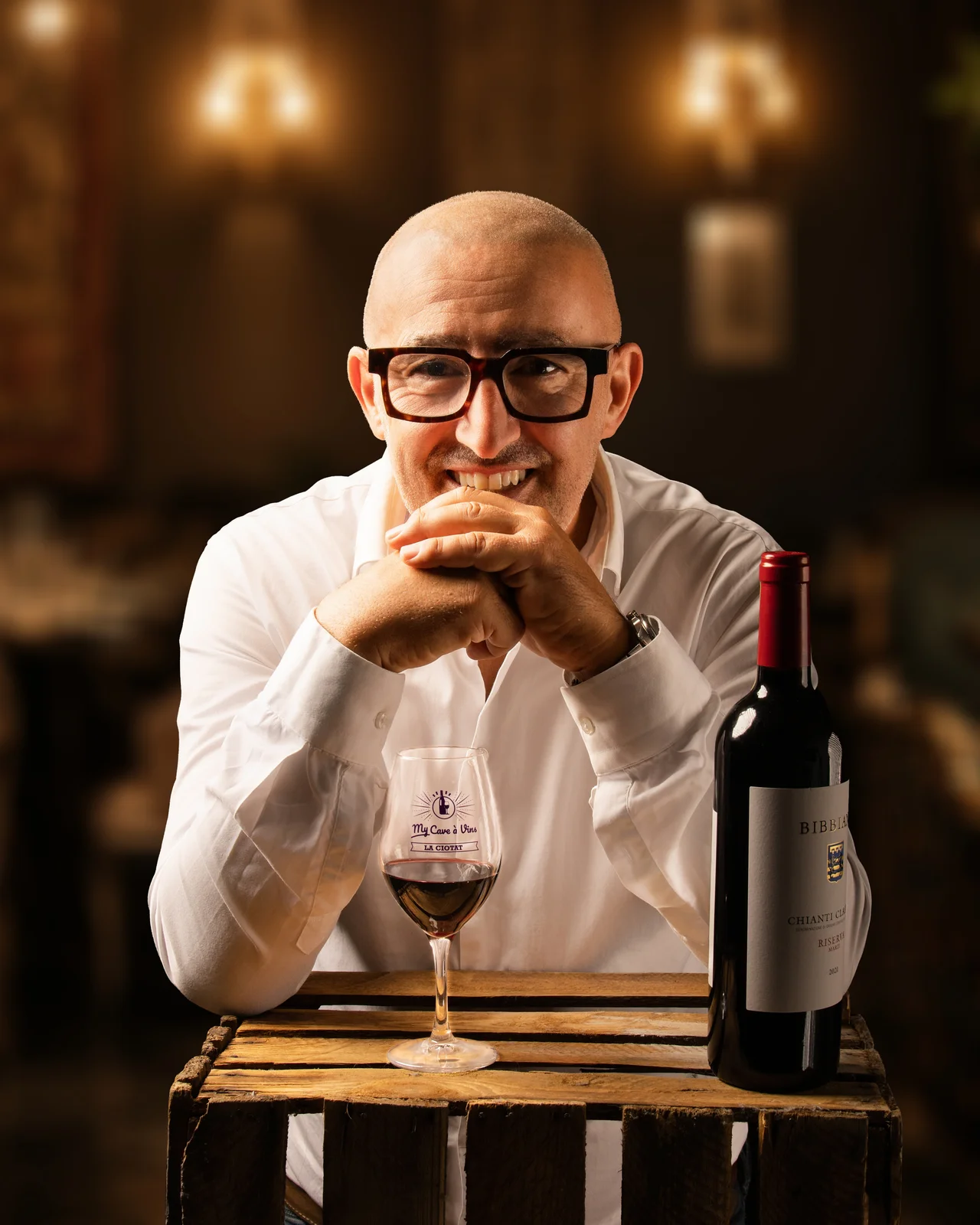 Homme chauve à lunettes, chemise blanche, mains jointes sous le menton, verre de vin rouge et bouteille sur une caisse en bois