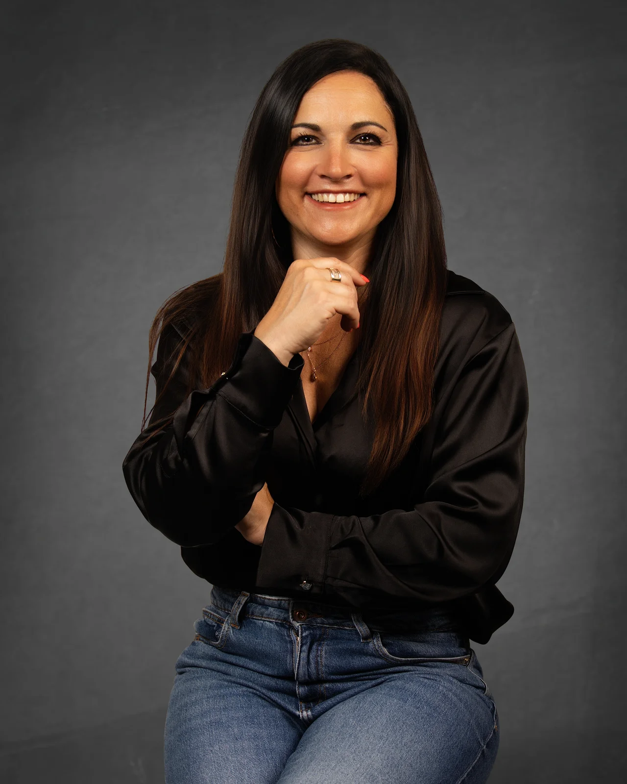 Femme brune souriante, chemise en satin noir et jean, main près du menton, assise sur fond gris en lumière douce de studio.