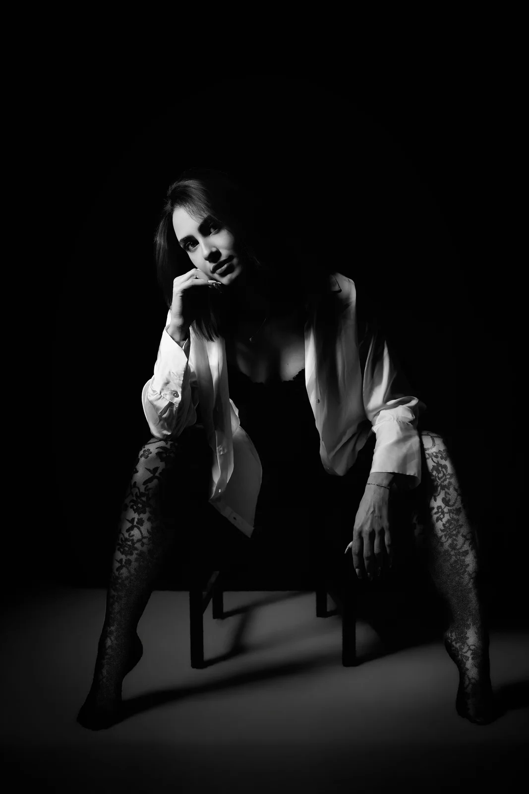 Femme assise sur un tabouret, chemise blanche ouverte et collants en dentelle, menton sur la main, noir et blanc en clair-obscur