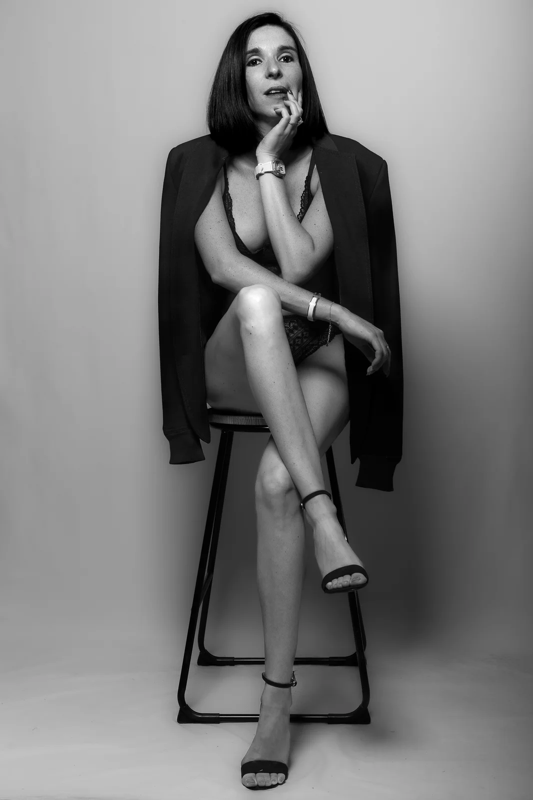 Femme brune en lingerie de dentelle, veste sur les épaules, assise jambes croisées sur un tabouret, noir et blanc en studio