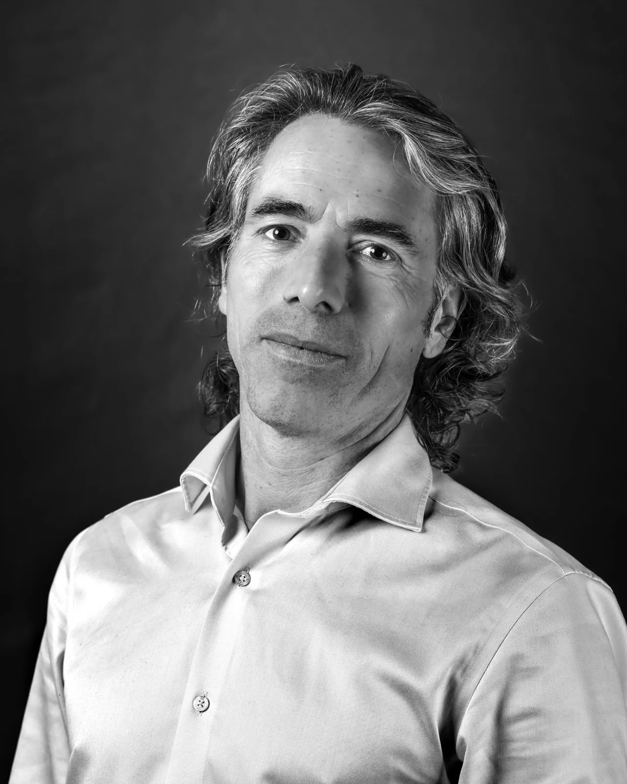 Portrait noir et blanc d'un homme aux cheveux mi-longs ondulés, chemise claire, léger sourire, sur fond sombre en studio.