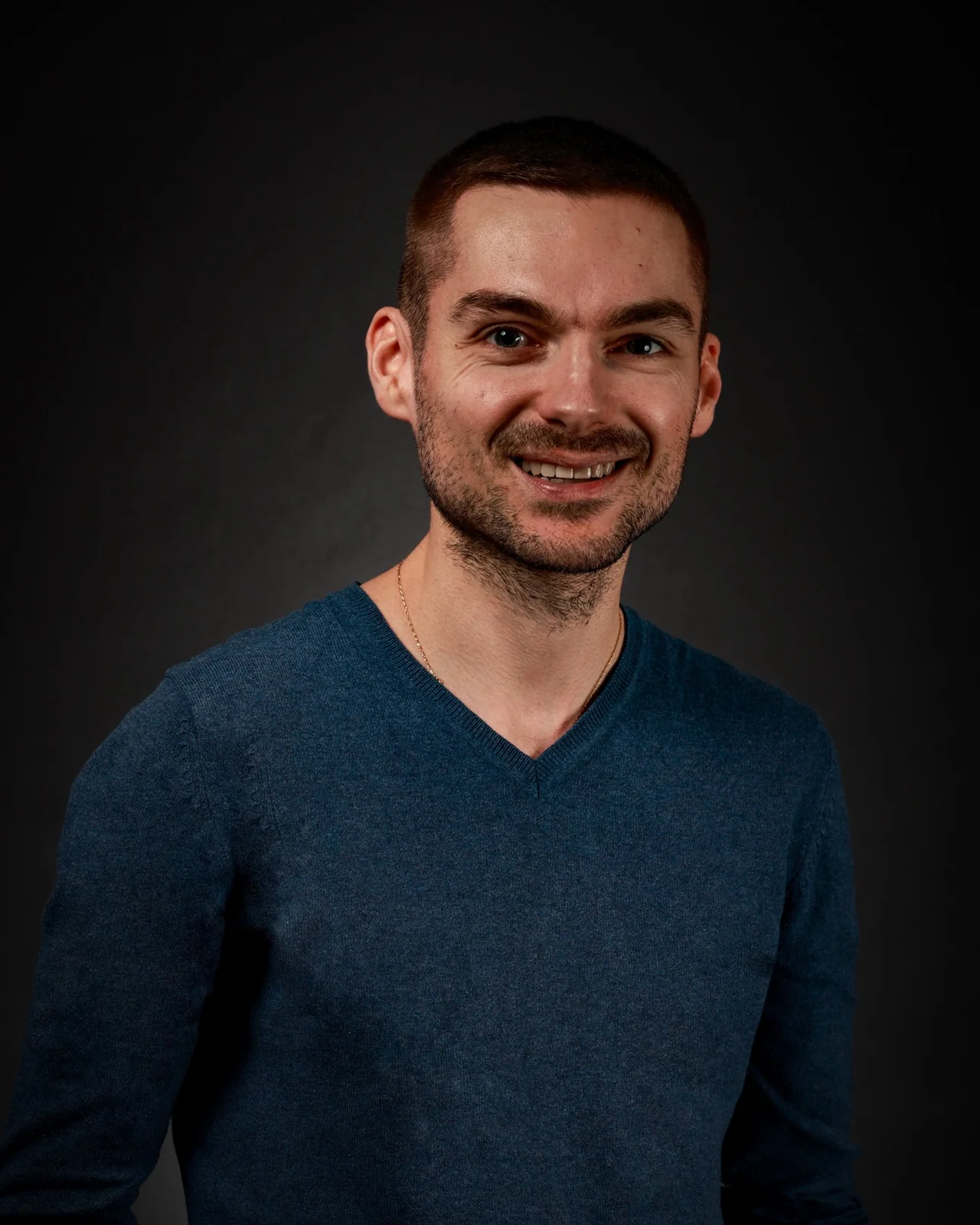Homme souriant aux cheveux courts et barbe naissante, pull bleu col V et chaîne fine, sur fond gris sombre, lumière douce.