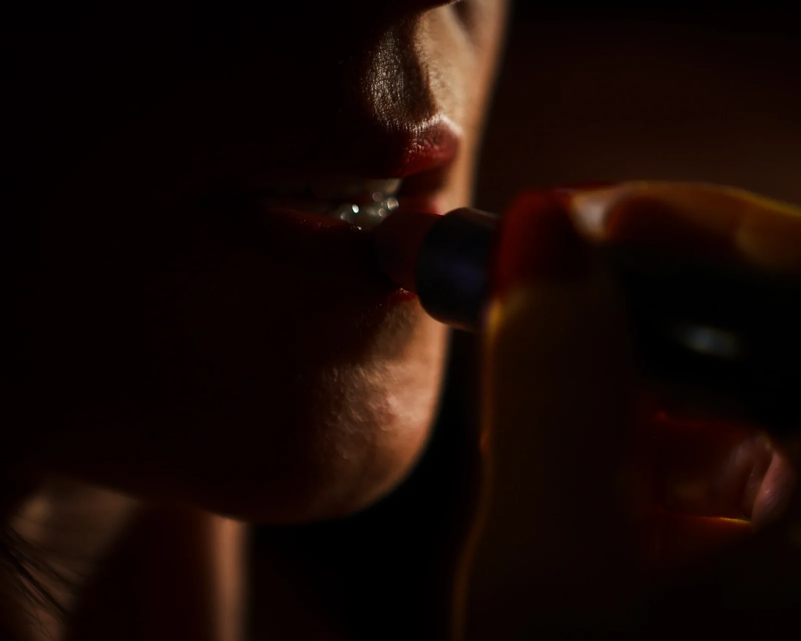 Gros plan en clair-obscur sur une bouche qui applique un rouge à lèvres rouge, main floue au premier plan, lumière chaude